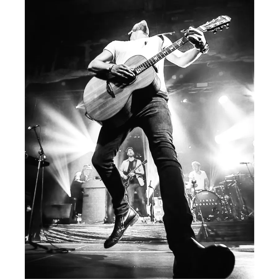 Connor McDonald mid-jump on stage with an acoustic guitar under stage lights